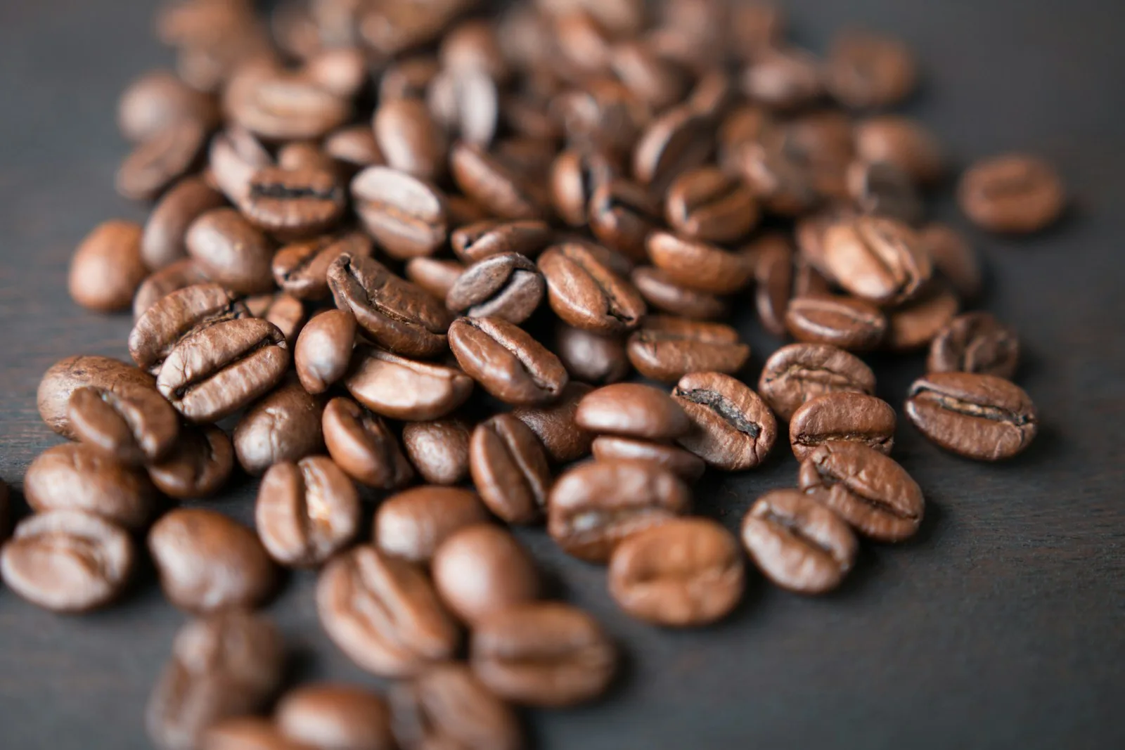 Macro of roasted coffee beans scattered on a dark wood counter, hand grinder and brass scoop beside them