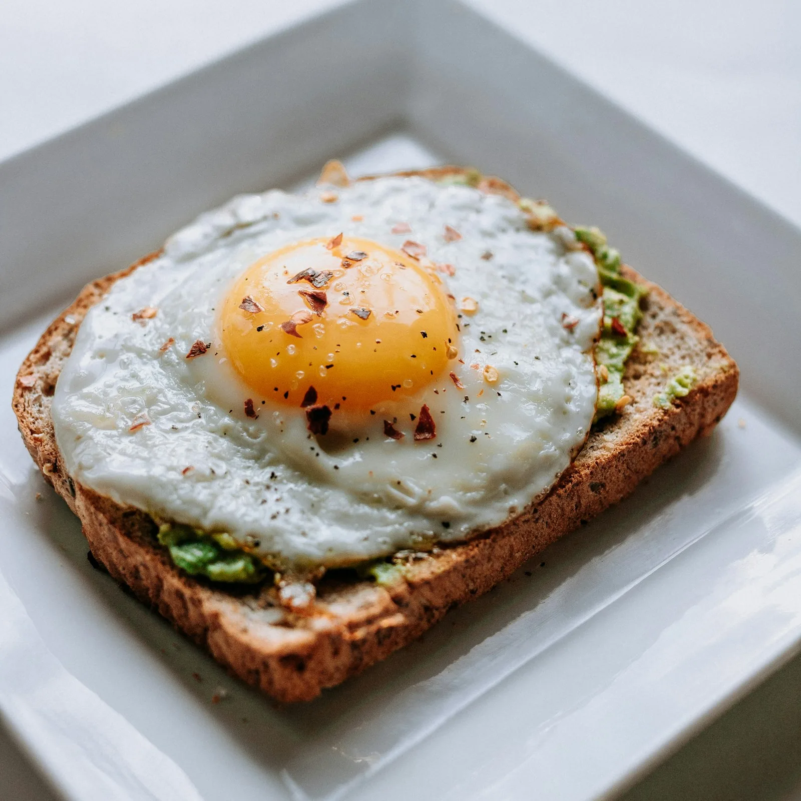 A square white plate with avocado toast and a soft-boiled egg cut in half, beside a small glass of orange juice, stone wall and green plants in the soft background