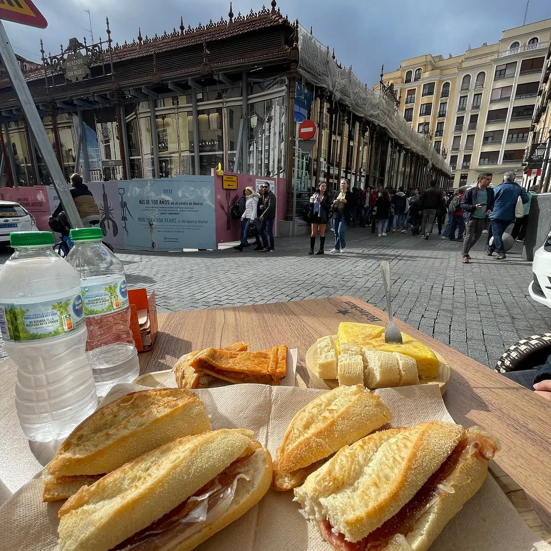 Two jamon bocadillos and a wedge of Spanish tortilla on a wooden table outside a covered market