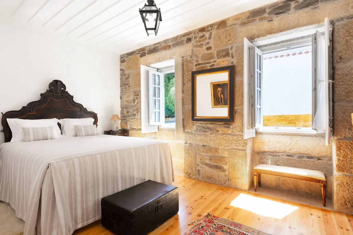 The Xisto room at first light: a long stone wall, a woodstove, a bed with linen sheets, two open windows onto the schist terrace.